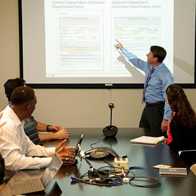 Business people gather around a desk with a man pointing at an image projected on a screen at the head of a conference table.