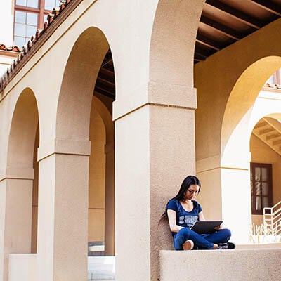 A woman sits under an archway at UC Riverside working on her laptop.