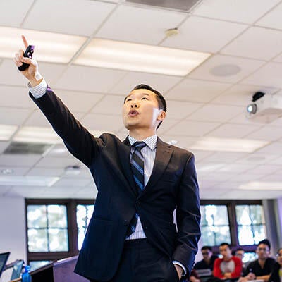 A man in a suit points at a screen (off camera) during a presentation.
