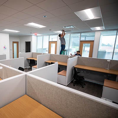 Two men work on infrastructure above drop ceiling in an office.