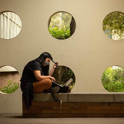 A woman sits on a short wall working on a computer.
